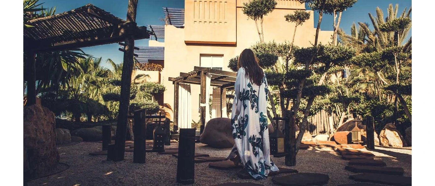 Elegant woman walking in a blue and white kaftan through a leafy courtyard towards a blush coloured stone building