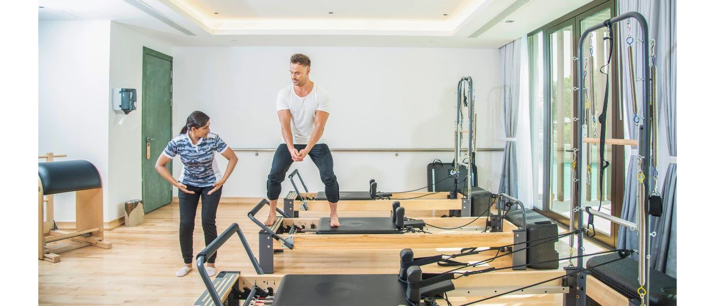 Man in white t shirt and black joggings bottoms works out on a Reformer Pilates machine as a woman next to him demonstrates