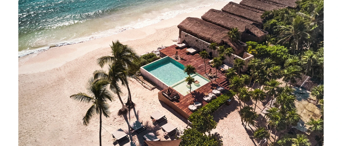 White-sand beach with turquoise shallows overlooked by a resort with thatched-roof buildings, an infinity pool and wooden deck covered in daybeds