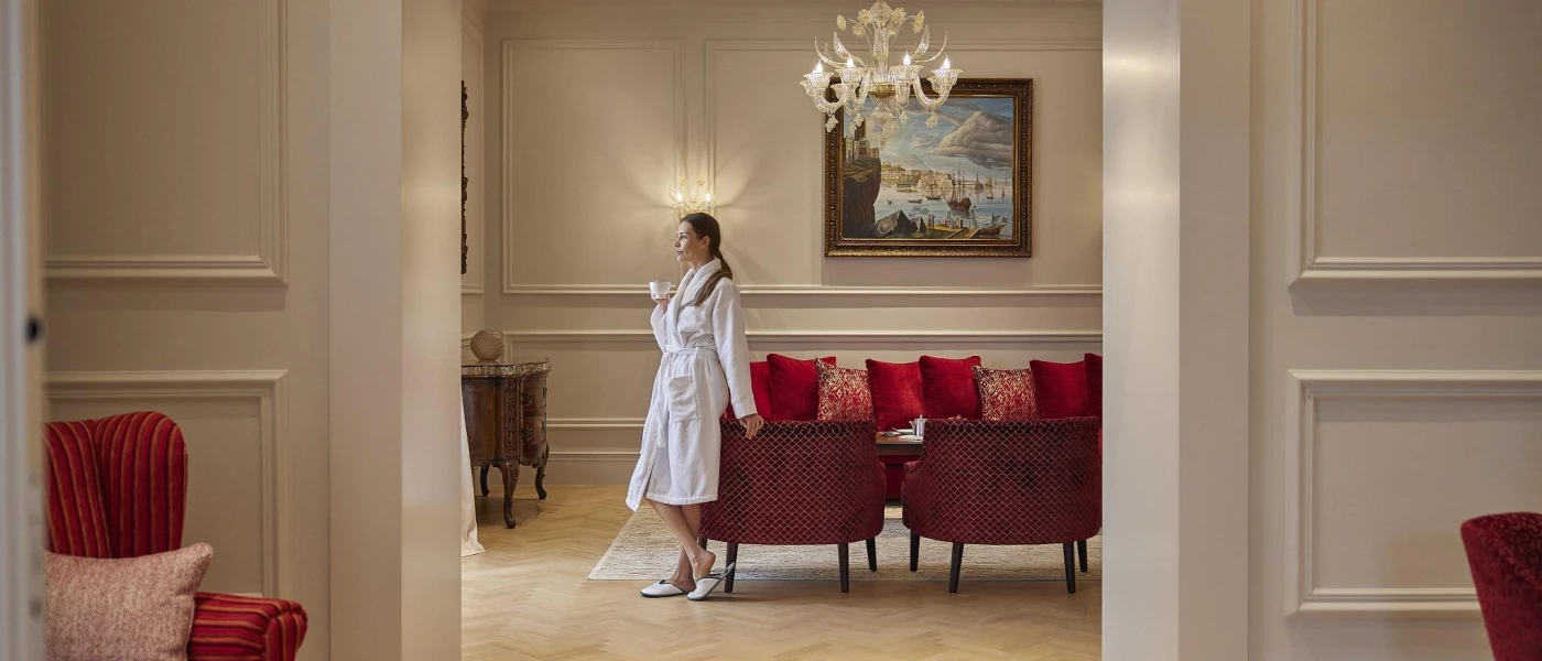 Woman in a white robe and slippers holding a china cup while leaning on a chair in a room with a chandelier and red-cushioned sofas