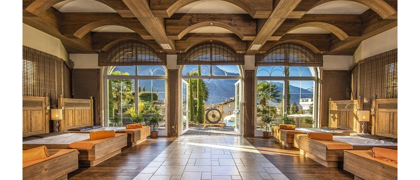Airy high ceiling wooden room with wooden daybeds with orange towels and blankets, looking onto a terrace with a large gong for sound healing and the surrounding mountains