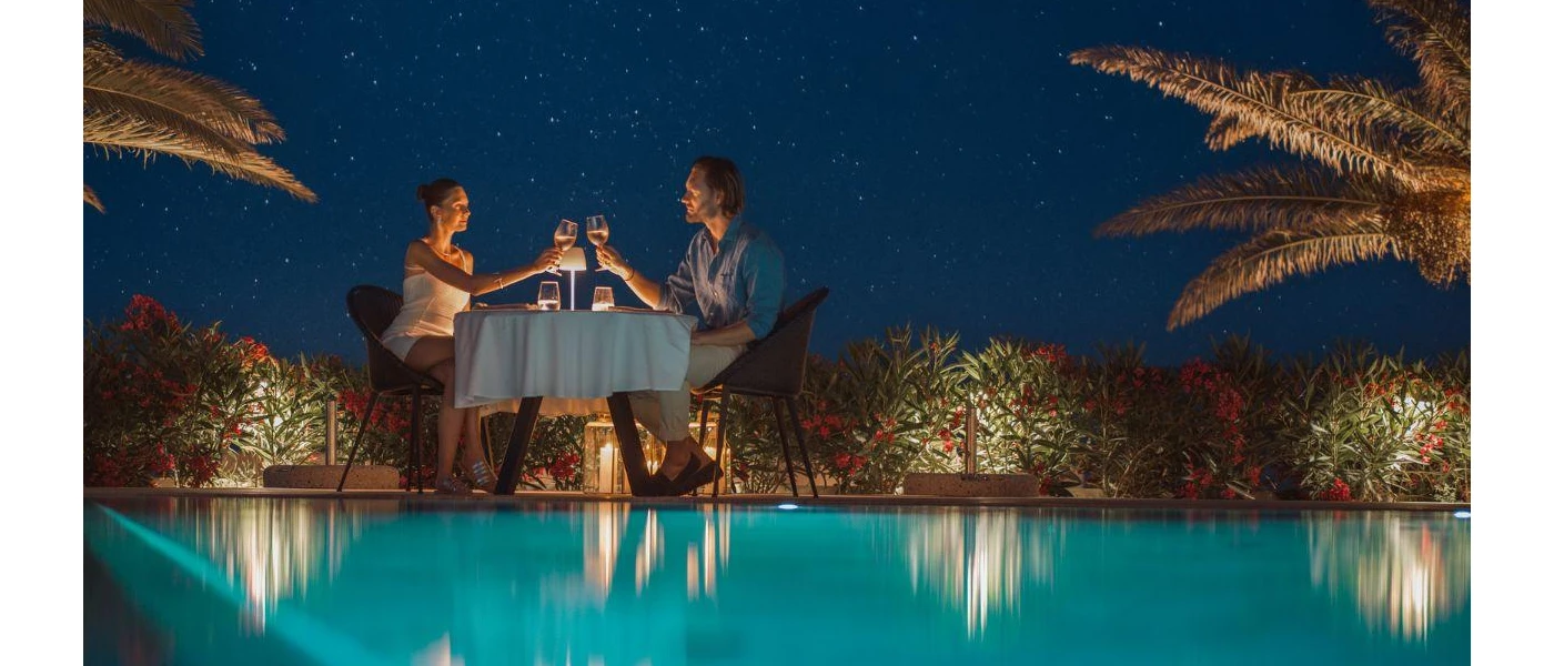 Couple toasts with wine glasses at a table set up by the poolside under a starry night sky