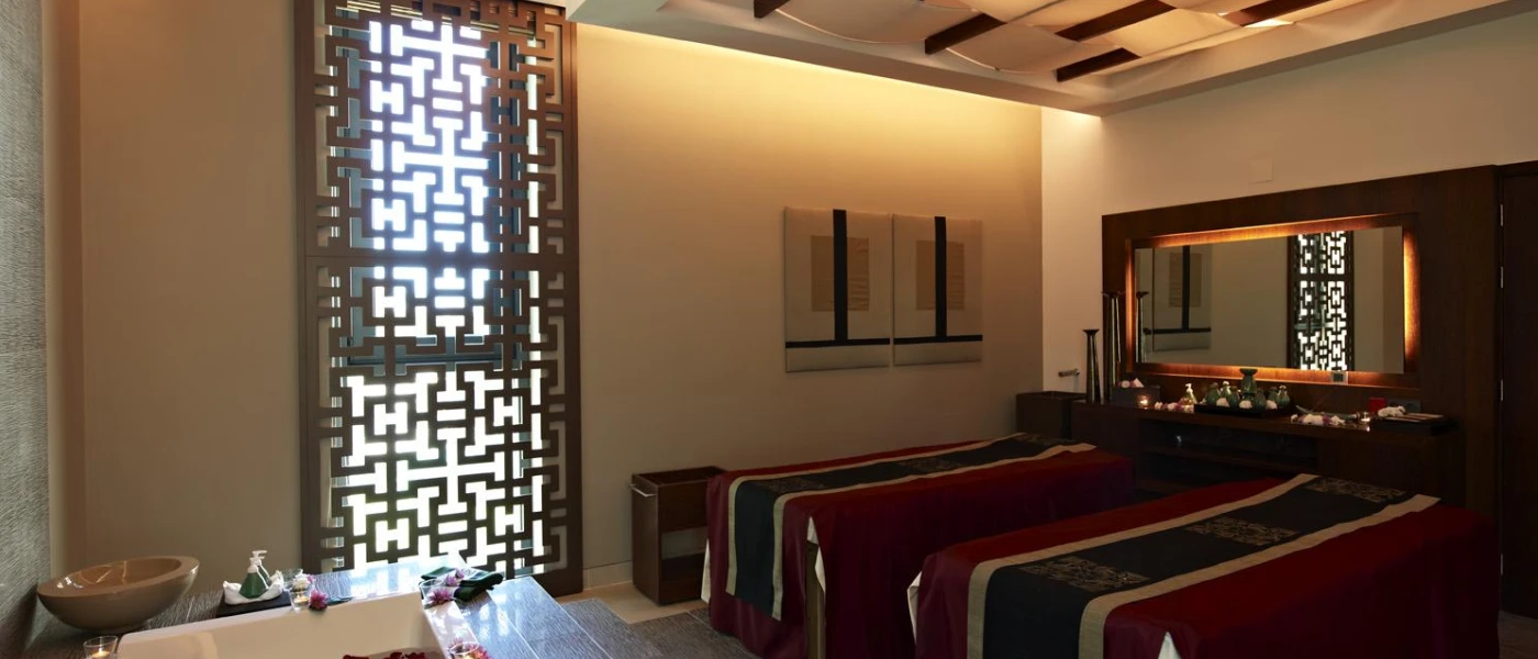 Cosy low-lit treatment room, with two beds dressed in red and black, a white bath filled with red rose petals, and a window covering carved in dark woods