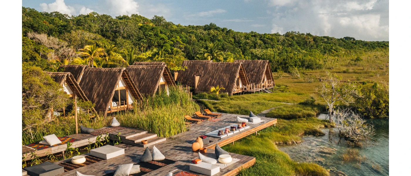 Rustic thatched-roof A-frame huts on a lakefront next to wooden decking and surrounded by jungle greenery