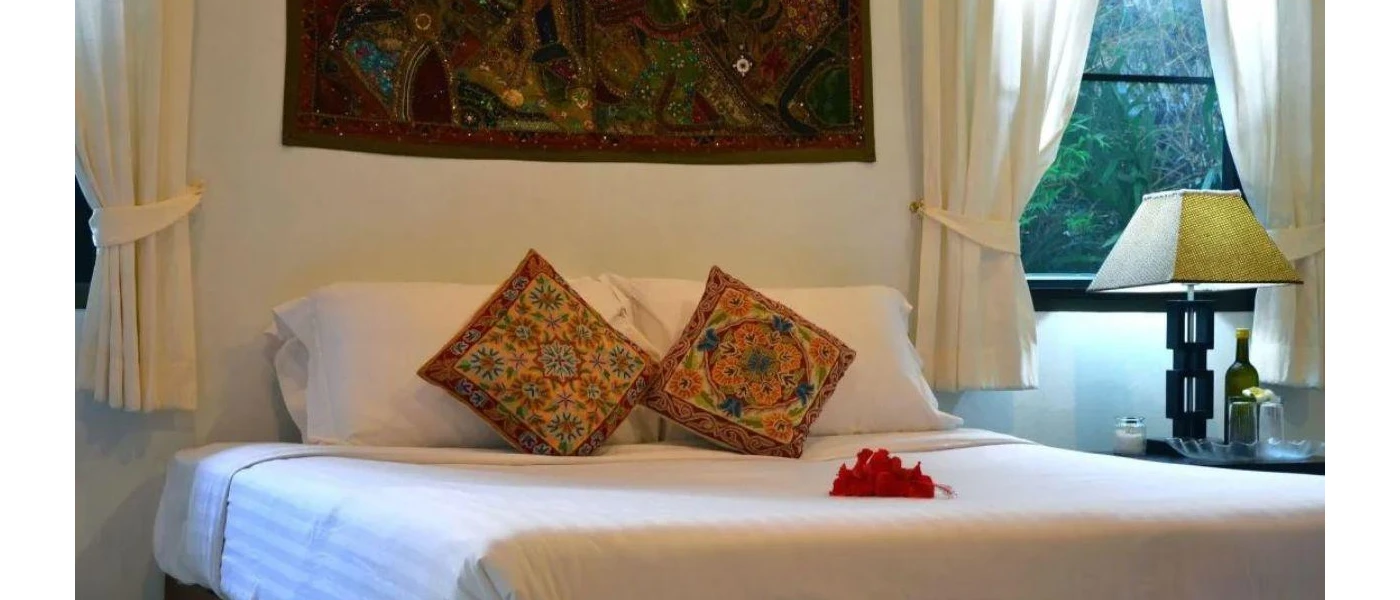 Bedroom with white bedding, patterned cushions, a window with a bedside table and lamp and a hanging material wall tapestry