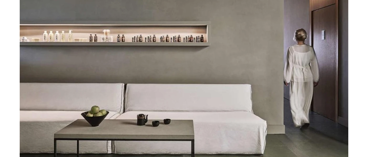 Woman in flowy white clothes walking out of a grey room with a white sofa and shelves full of lotions and potions