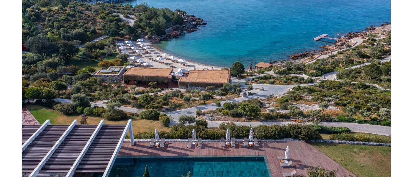 Hillside swimming pool set in wooden decking, with a view of greenery, a sandy beach and the Aegean Sea