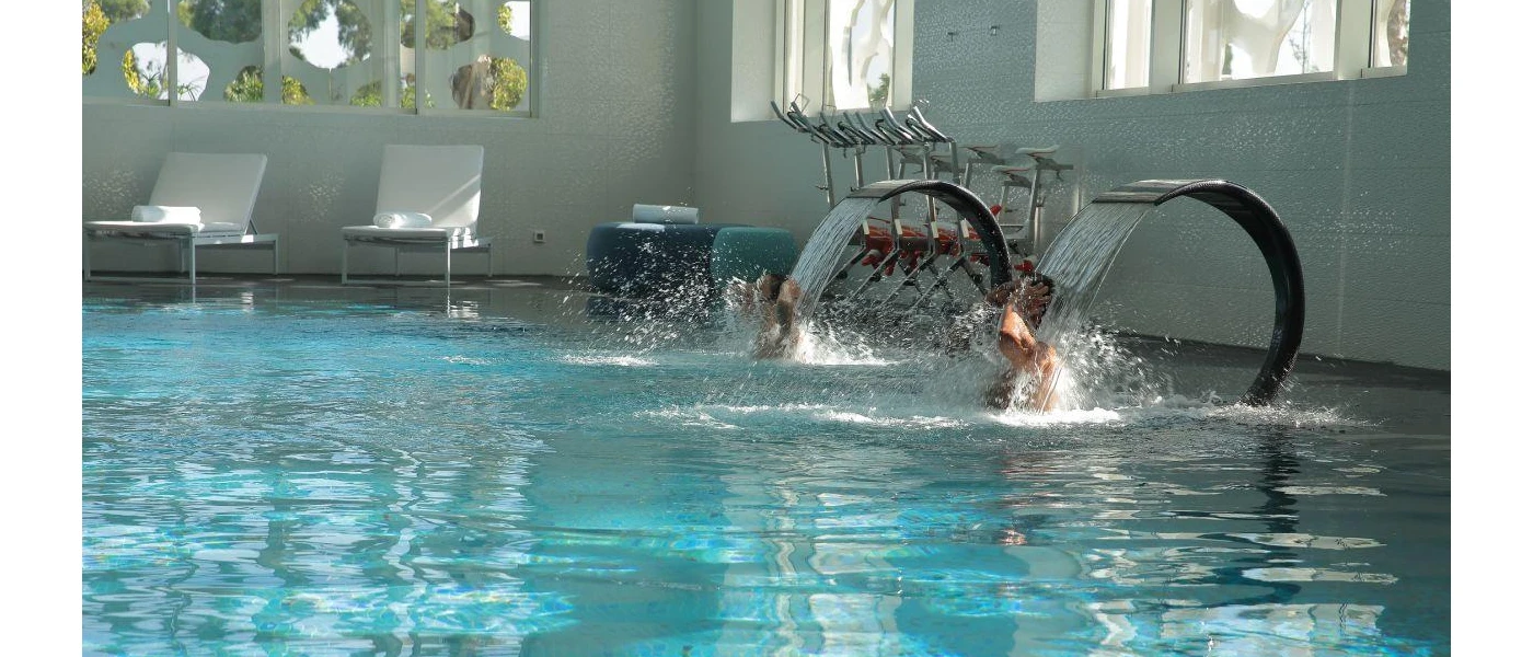 Two people enjoying the water jets at an indoor pool with two loungers in the background
