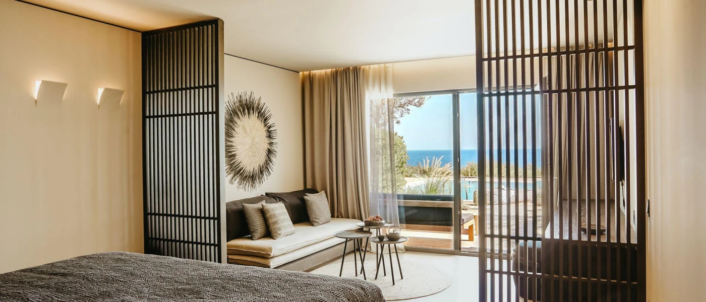 Hotel bedroom and living area with a bed with a charcoal fleecy throw, black partitions, cream walls, a sofa and cushions in neutral shades and a balcony with a sea view