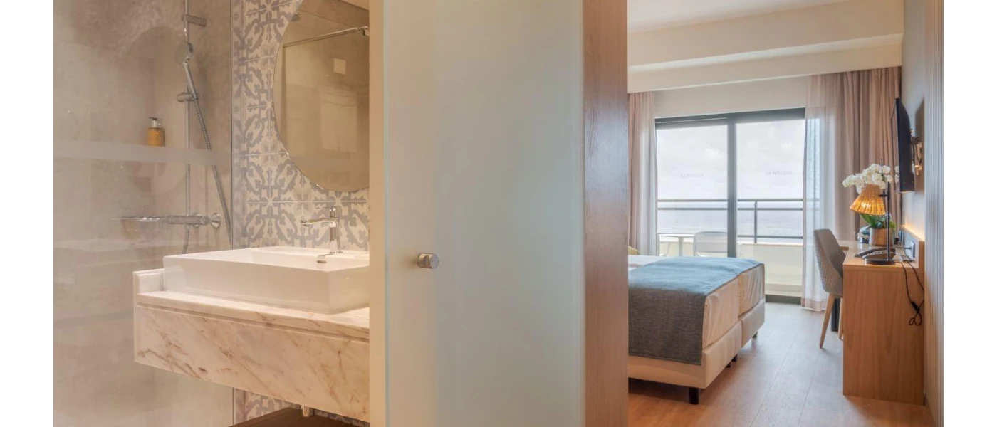 Partial view of a marble bathroom and the end of a bed with a blue throw, plus a balcony with an ocean view