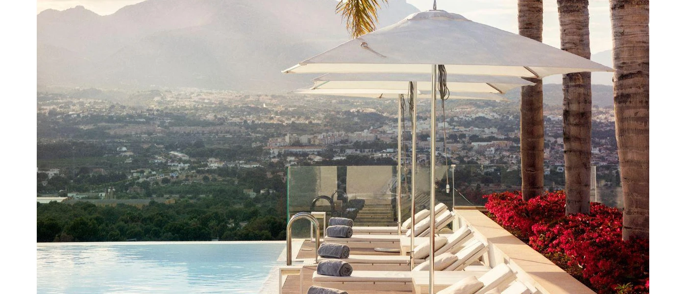 Infinity edge pool overlooking mountains and whitewashed buildings below, with sleek white-cushioned loungers and parasols topped with blue towels, and tall palm trees behind them