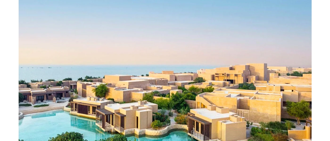 Low-rise, cube-shaped sand-coloured buildings huddled around a swimming pool under a blue sky with the sea in the background