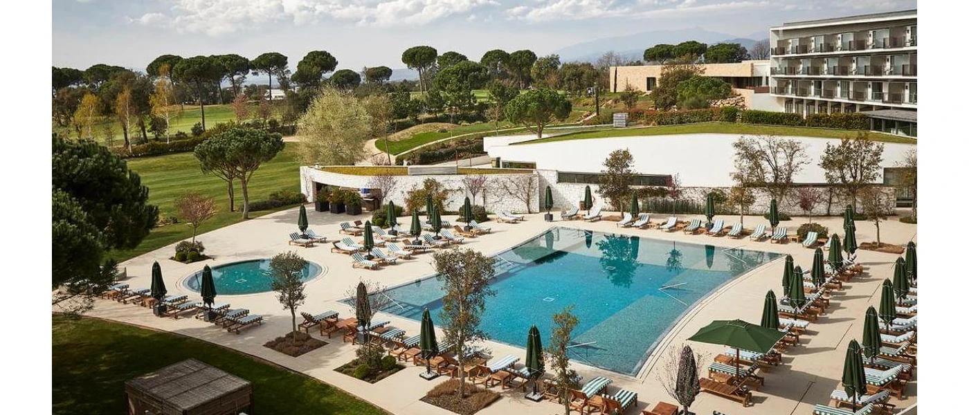 Stone terrace with a large rectangular pool and smaller circular pool, wooden loungers with green and white striped cushions and green parasols, surrounded by manicured lawns and trees
