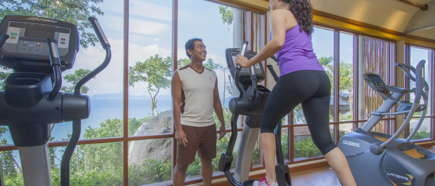 Woman in a purple vest and black leggings works out with a personal trainer in a gym with floor-to-ceiling windows