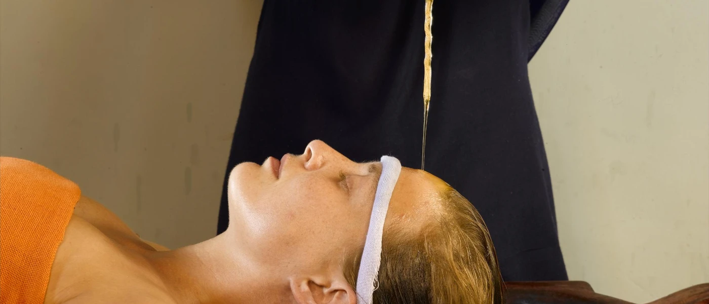 Woman lies on her back in an orange towel as a therapist pours oil over her forehead in a traditional Ayurveda treatment
