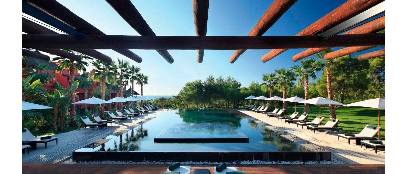 Rectangular raised swimming pool surrounded by cushioned loungers and white parasols and tropical greenery