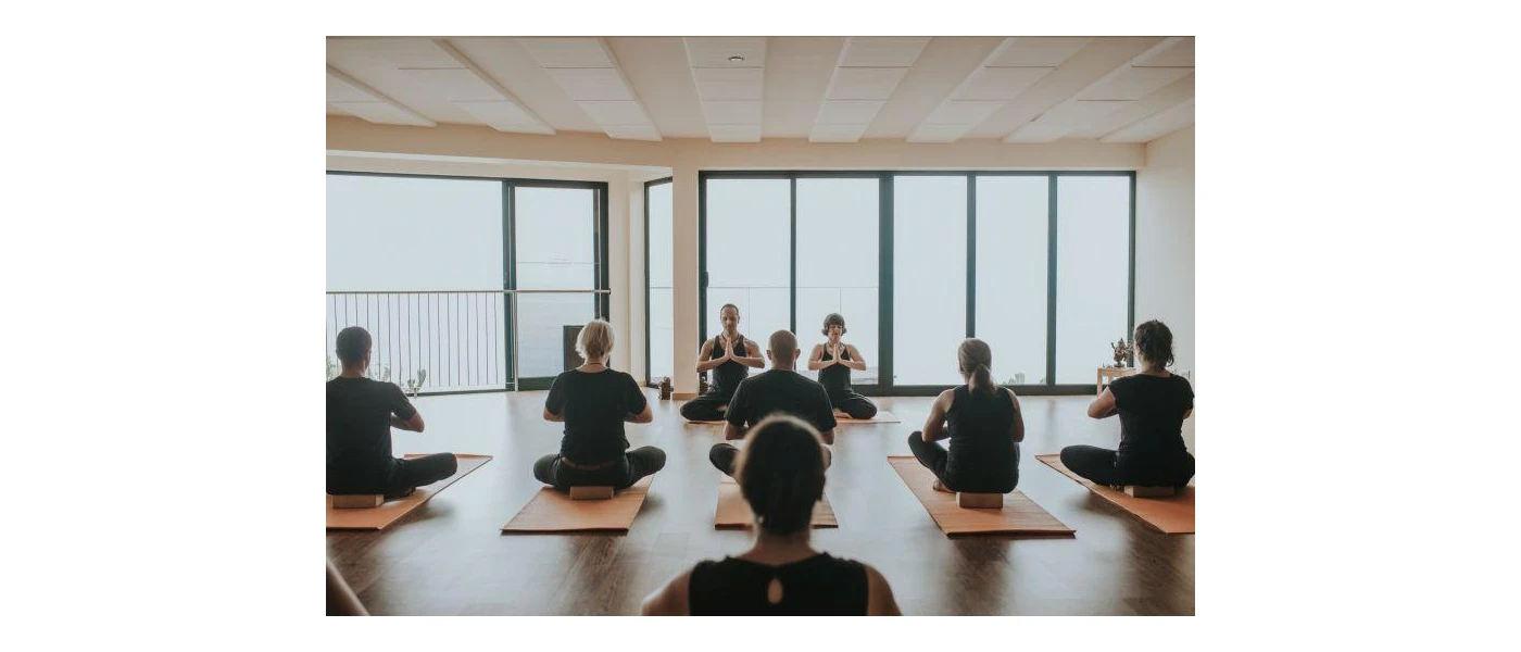 Group of people wearing black sit on blocks on yoga mats in a studio with floor-to-ceiling windows
