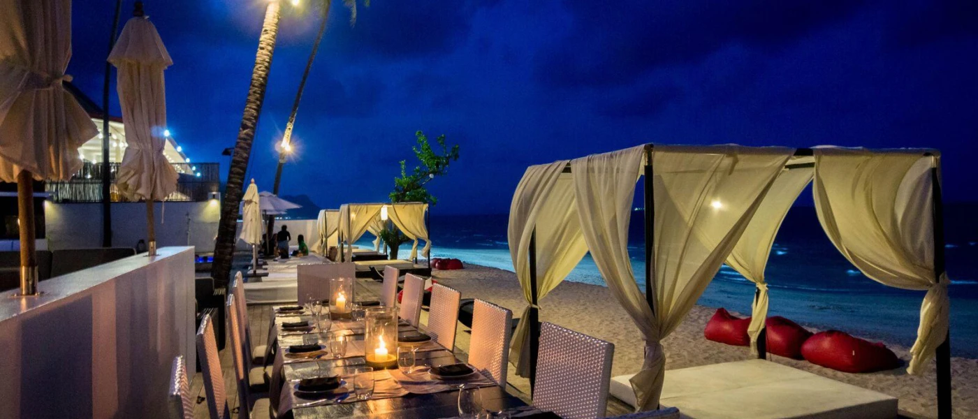 Candelit tables on a beachfront terrace, fronted by a four-poster cabana and daybed draped in white fabric