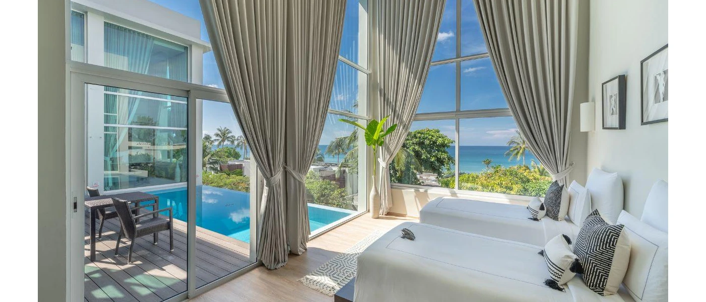 Airy room with high ceilings, white walls and bedding, window drapes and floor-to-ceiling glass overlooking a pool terrace and the ocean