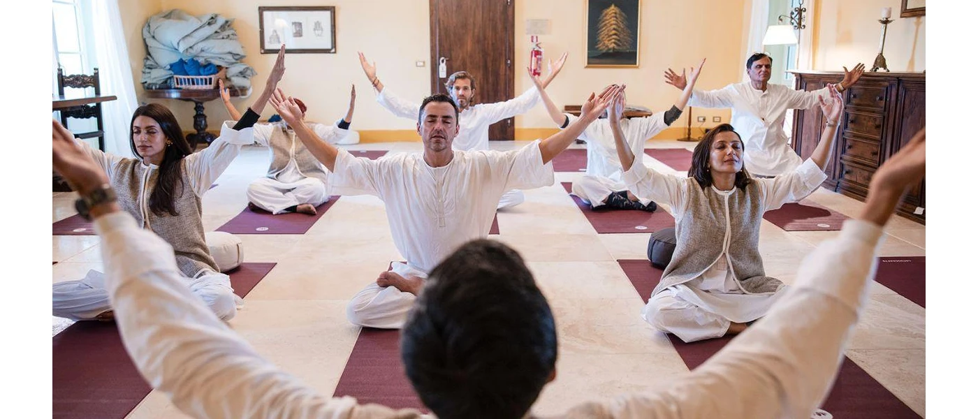 Group in loose white clothes sit cross legged with arms outstretched and eyes closed on purple yoga mats