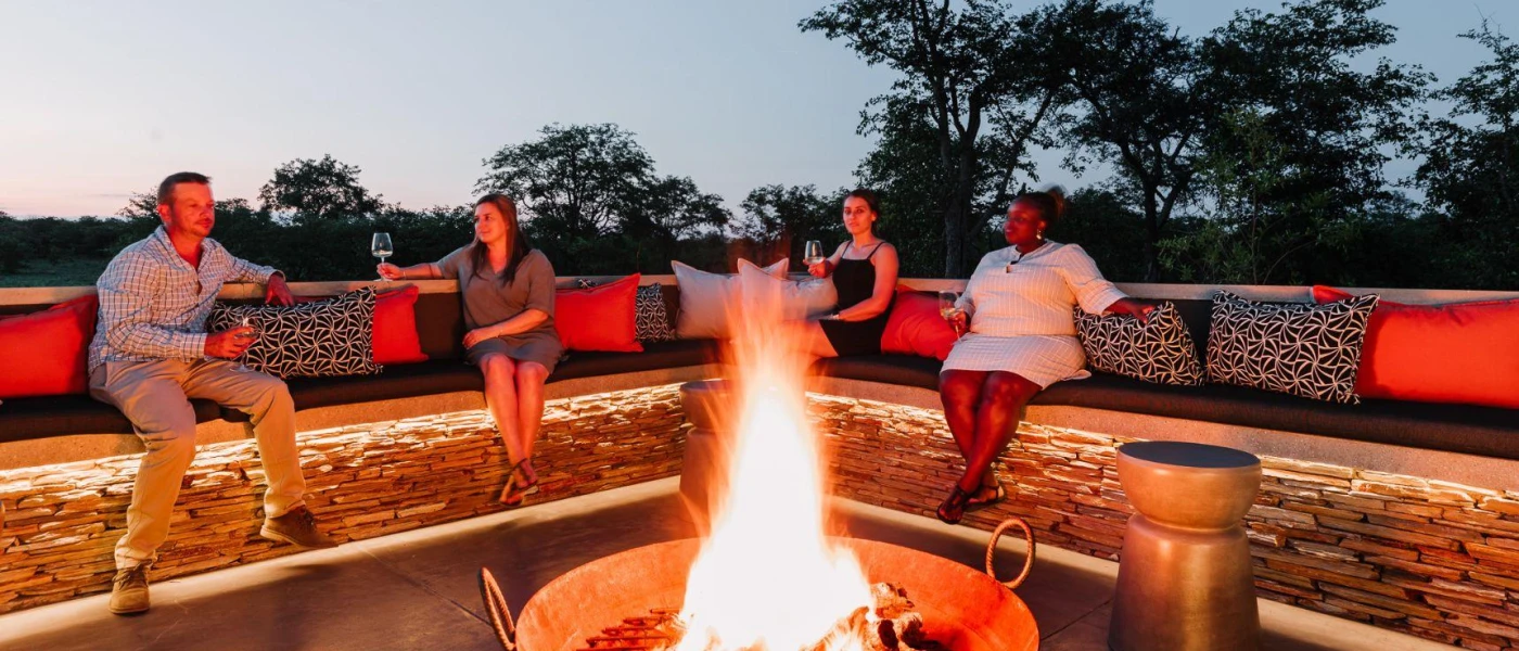 Guests relax with glasses of white wine around a central fire pit under a darkening sky