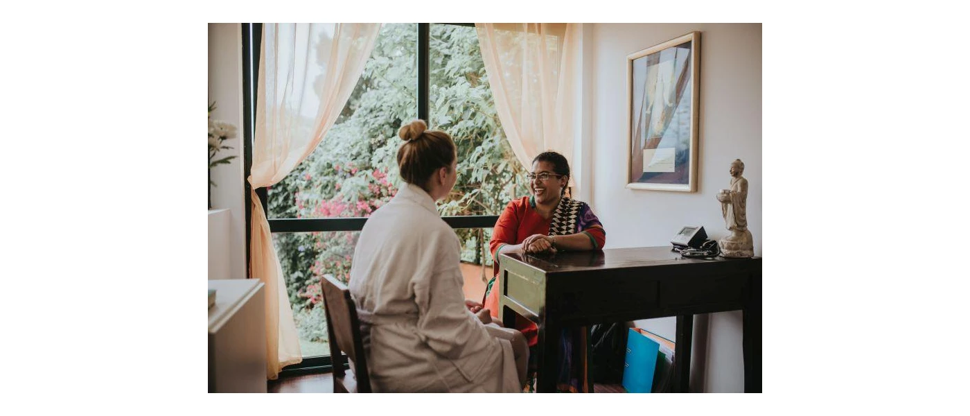 Woman in a robe sits at a wooden desk with a smiling Ayurveda expert