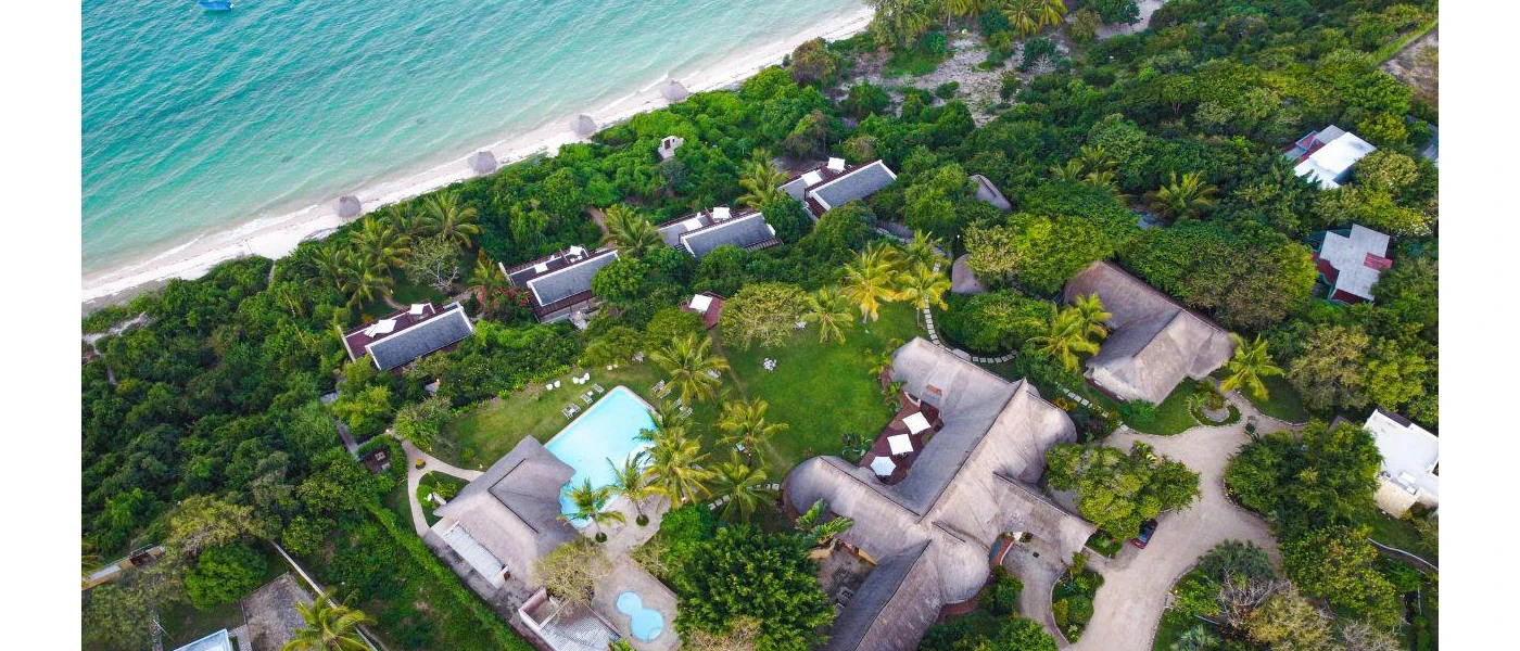 Aerial view of tropical greenery, grey-roofed buildings and a white-sand beach and ocean