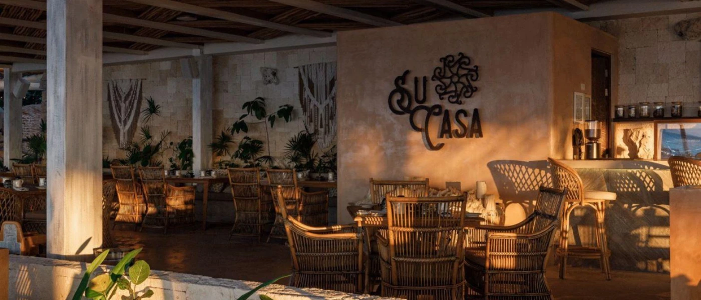 Rustic restaurant with beige walls, rattan chairs, macrame designs and plants, and the words 'Su Casa' etched on the wall