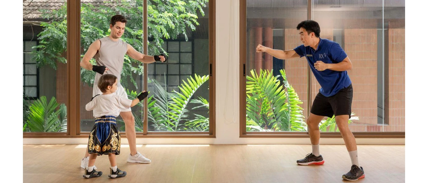 A man in active wear and a boy in Thai boxing shorts practice boxing with a staff member in a blue polo shirt