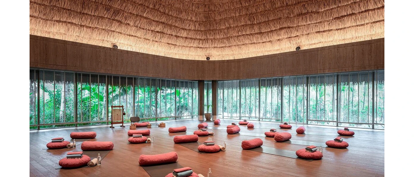 Thatched-roof pavilion with floor-to-ceiling windows, set up for a class with grey yoga mats, red cushions, blankets and bottles of water, with a gong at the head of the class