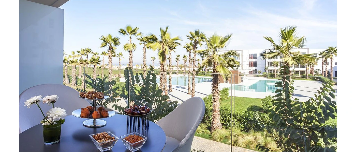 Balcony table set up with fruit overlooking a swimming pool surrounded by palm trees and lawns