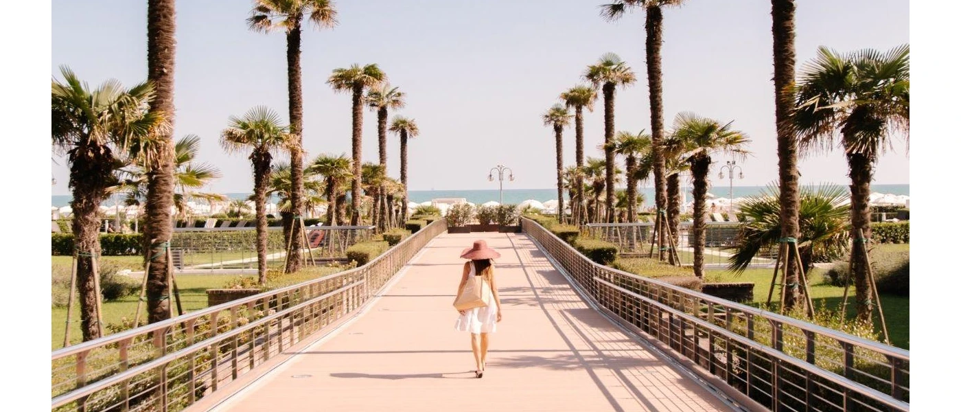 Woman in white dress and straw hat strolls across a paved bridge lined by palms towards the sea