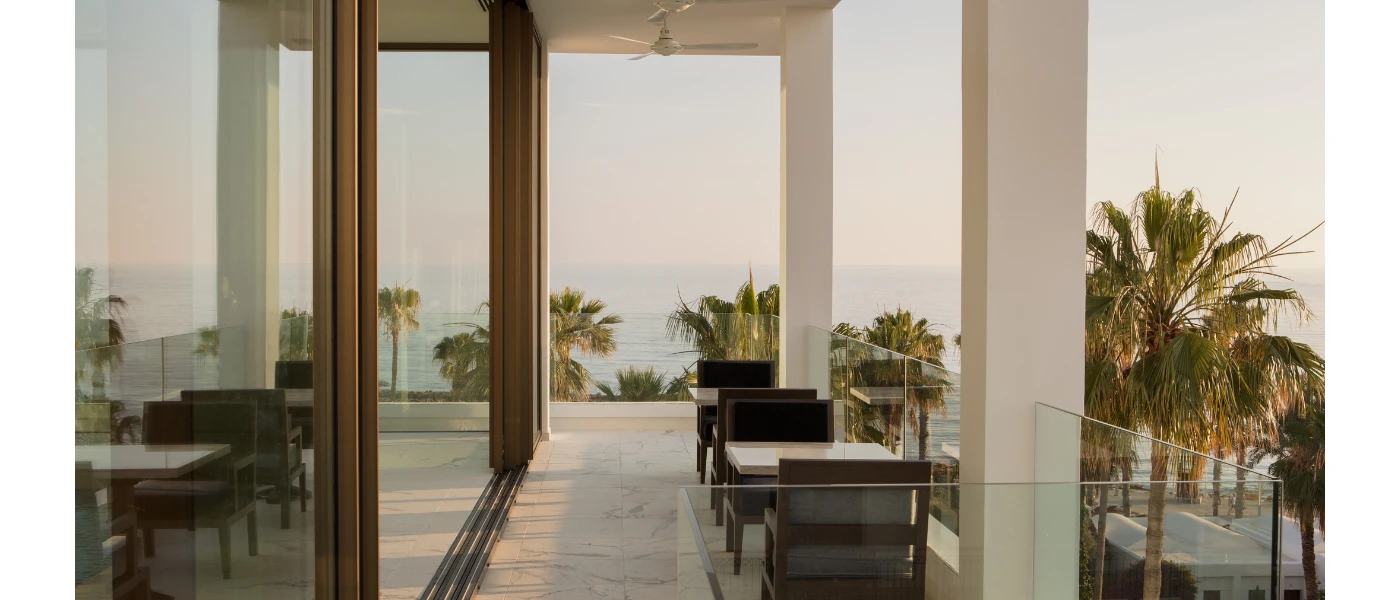 Glass-fronted balcony with chairs and tables, overlooking the sea in the distance
