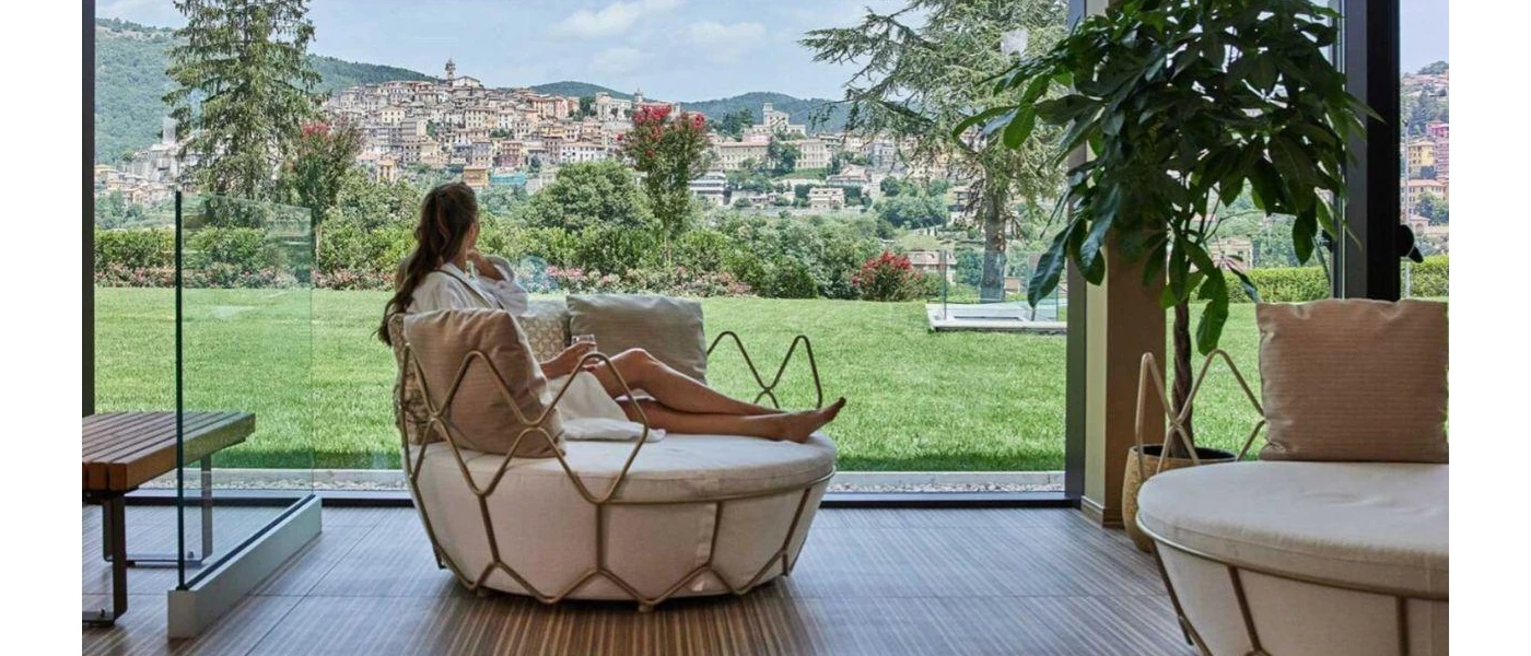 Woman in a robe sitting in a cushioned round chair, overlooking a lawned garden and the hillside town of Fiuggi