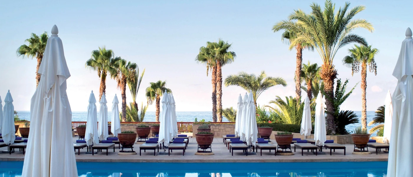Swimming pool lined with white parasols, white-cushioned loungers with blue rolled towels on top, tall palm trees and a view of the sea 