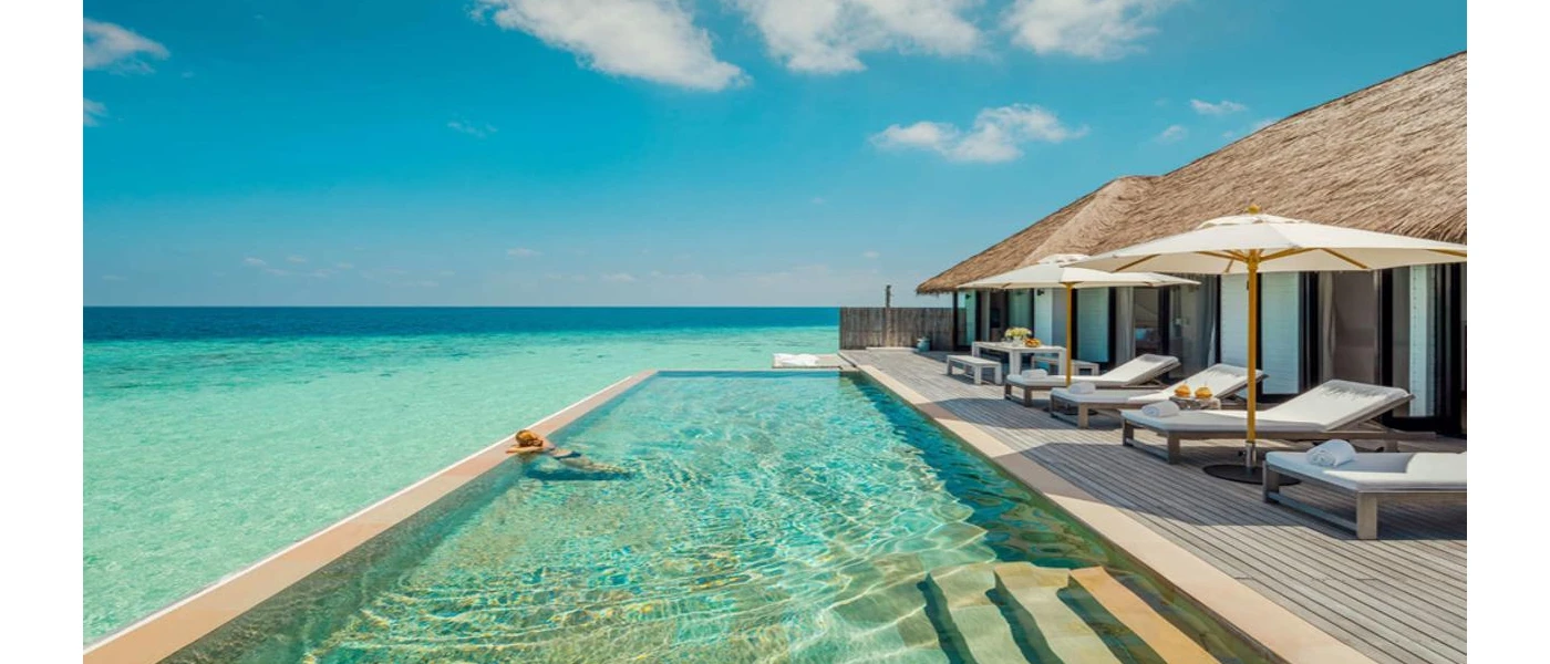 Woman relaxes in an infinity pool in a wooden deck furnished with white-cushioned loungers and umbrellas surrounded by turquoise shallows 