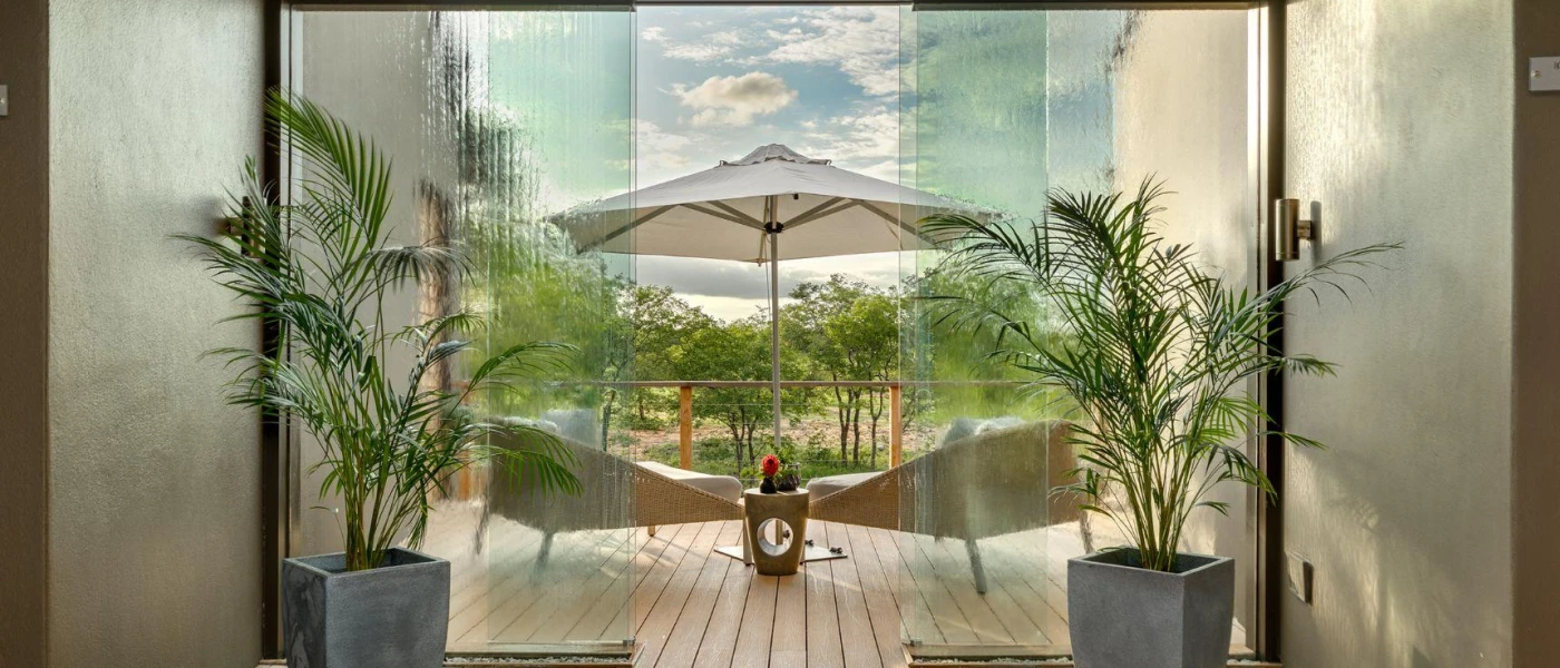 Wood-decked balcony with chairs and a white parasol, a glass door entry and two potted plants 