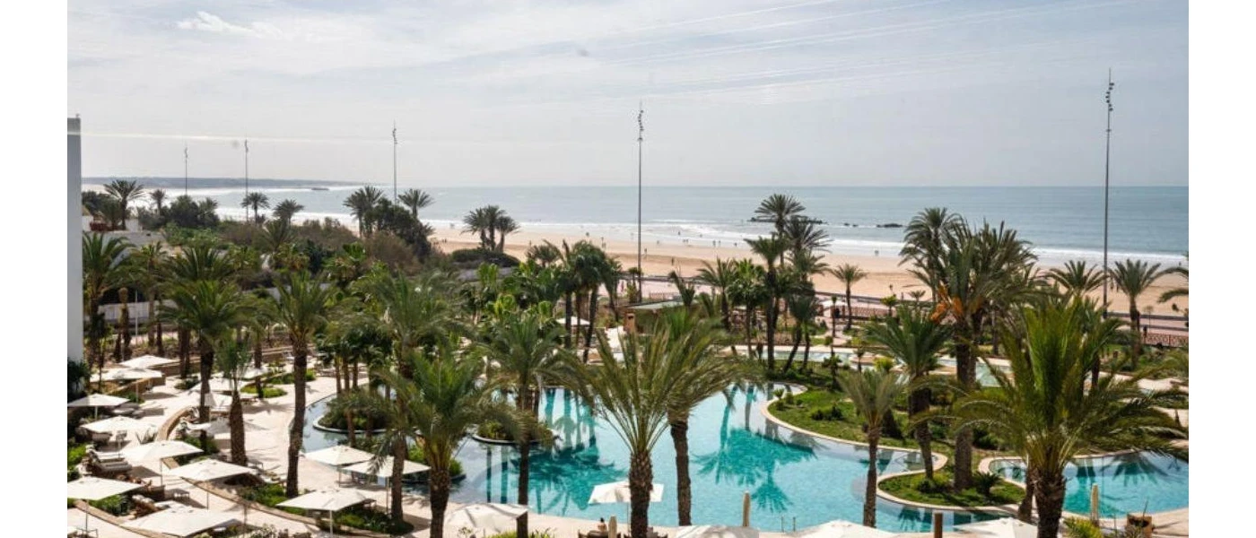 Palm trees and white parasols surrounding a swimming pool with the beach and ocean in the background