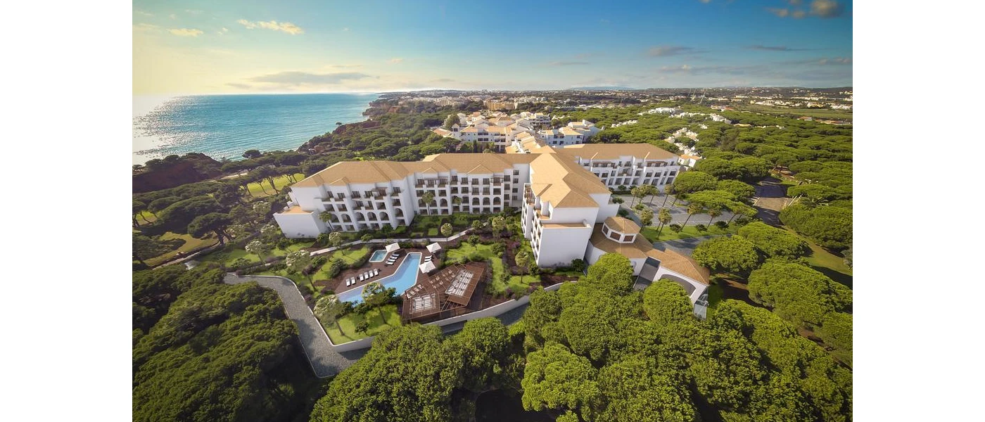 Multi-storey white building with light tiled rooftops, spread among leafy grounds with a pool next to the ocean
