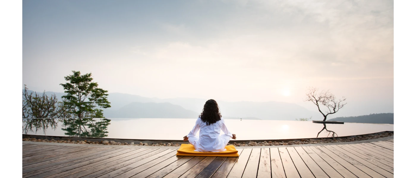 Woman sits on the edge of an infinity pool on a wooden deck meditating with misty hills in the distance