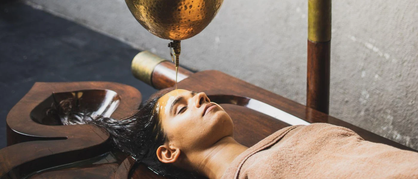 Woman lies on a dark-wood bed as an unseen therapist pours oil on her head from a gold bowl