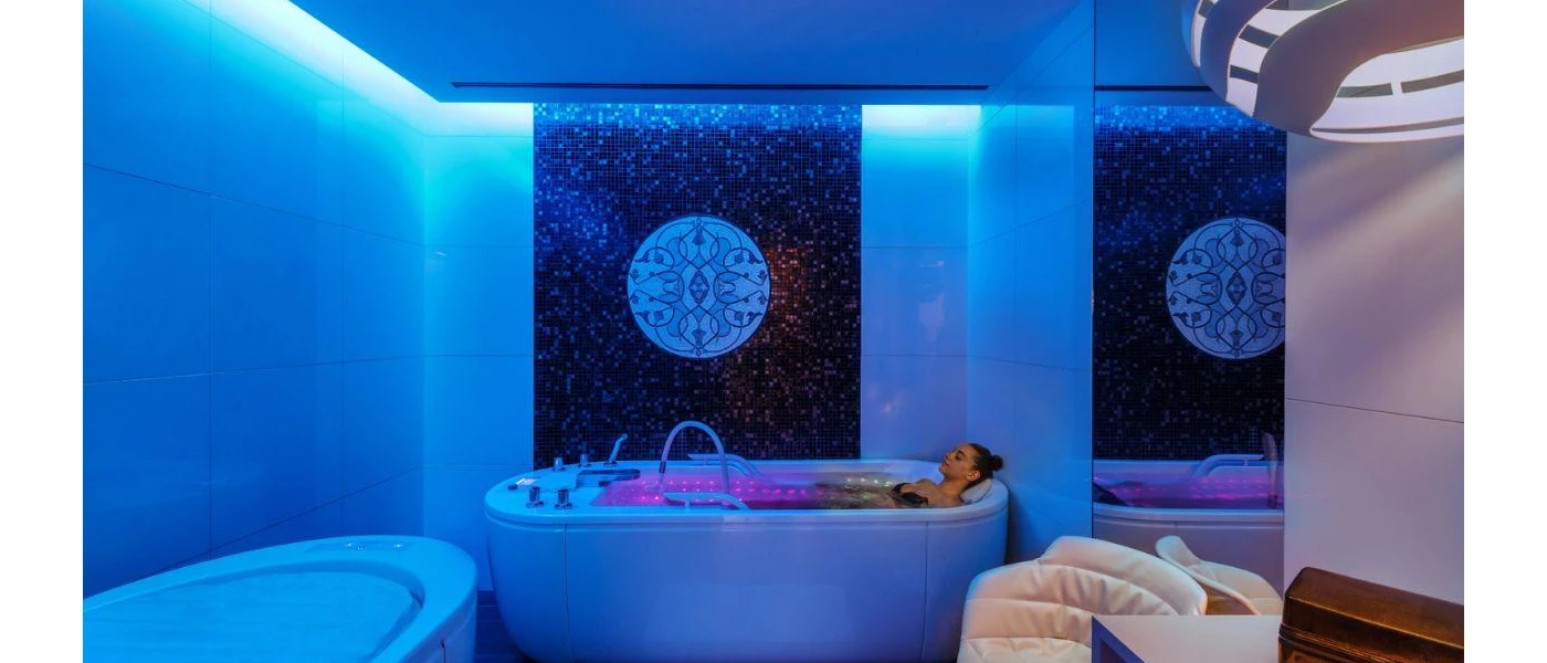 Woman in black swimwear relaxing in a filled bathtub in a white room with atmospheric blue mood lighting
