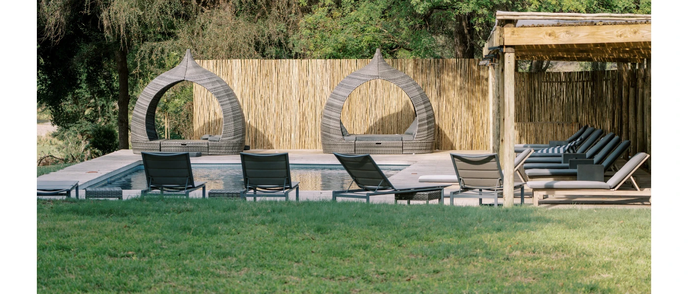Rectangular swimming pool with striped cushion loungers and a wooden canopy, and rattan domed shelters next to a grassy lawn