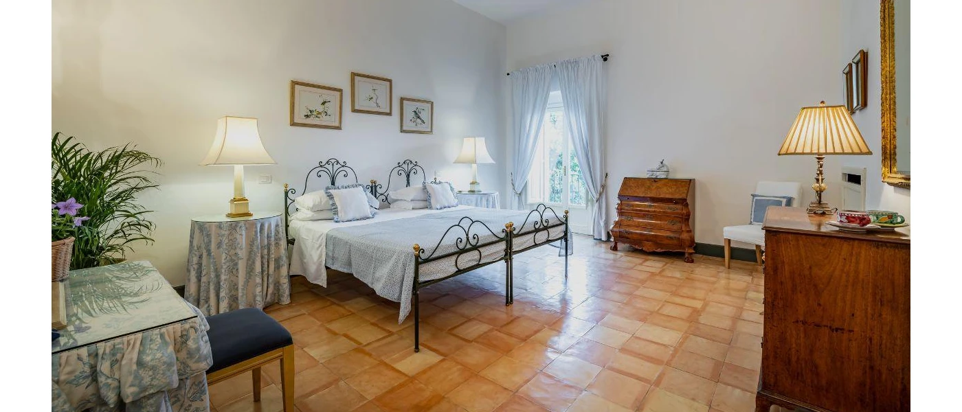 Traditional bedroom with a black iron bed frame, dark-wood furniture, tables with floral blue and white coverings and framed pictures on the white walls