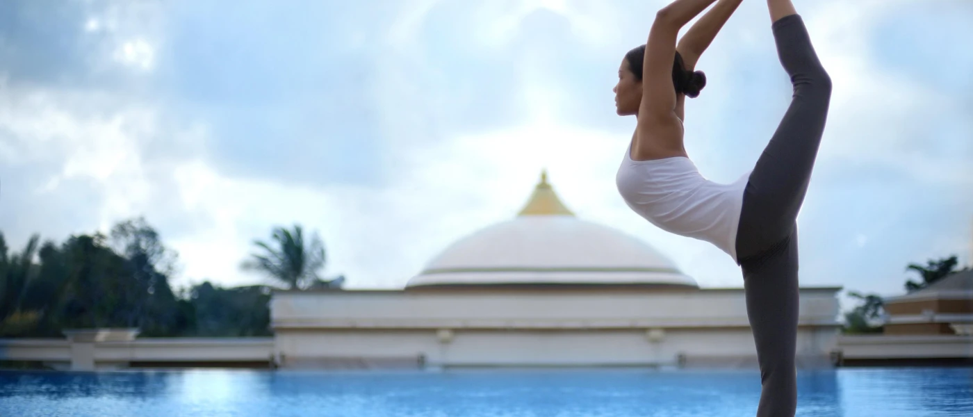 Woman lifts her leg high into the air next to a large swimming pool under a blue cloudy sky