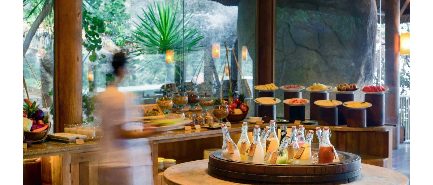 Buffet stations with plates of fruit, glass bowls filled with cereals and a central barrel filled with glass bottles with various drinks inside