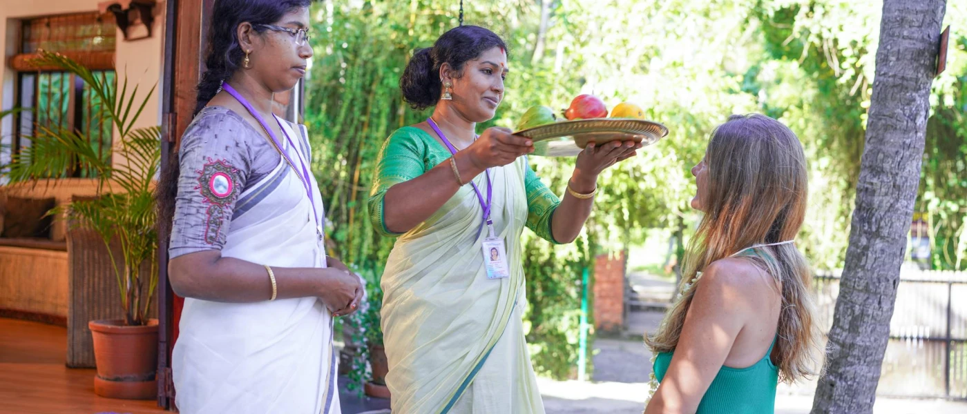 Two staff members in traditional dress stand in front of a woman wearing a floral necklace, as one woman holds a tray of vegetables up