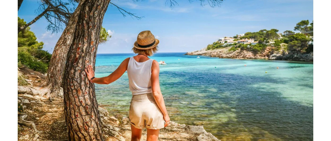 Woman in a white vest and shorts in a straw hat leans against a tree looking at turquoise waters