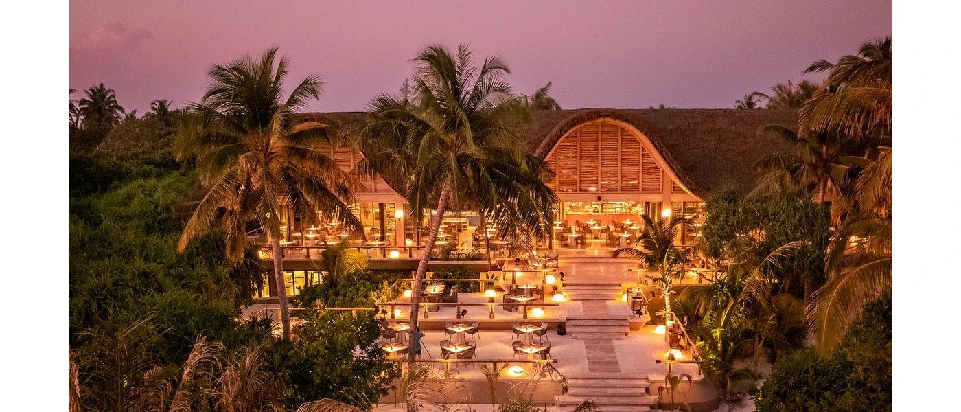 Stepped terraces down to the beach, cloaked in palm trees and shrubs, with tables and chairs covering the terraces and thatched rooftops in the background