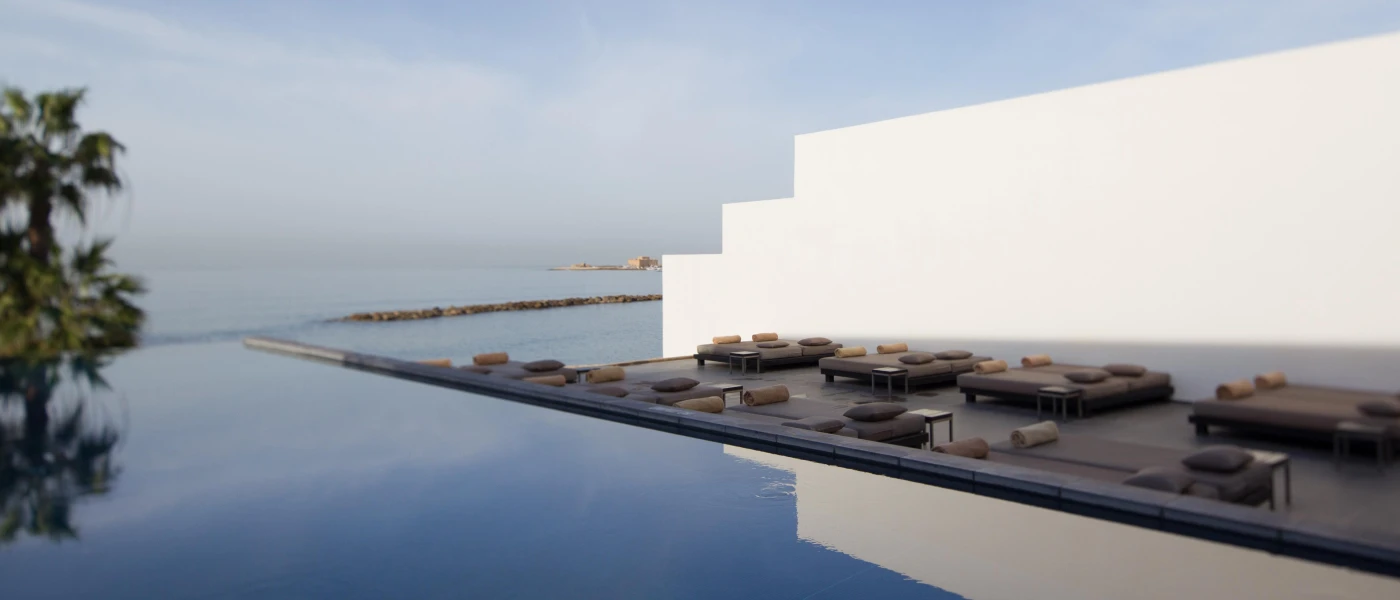 Edge of an infinity pool looking over palm trees and the ocean, with a line-up of cushioned sun loungers on the neighbouring terrace